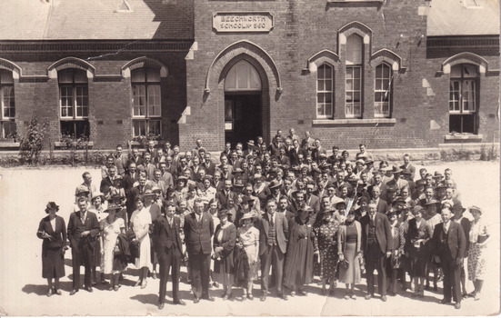 A sepia rectangular postcard with a large crowd of men and women standing in the foreground. Behind them is the Beechworth School building.
