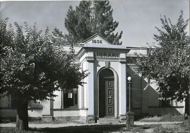 Two medium sized trees stand either side of the entrance to a classical style building, above the doorway is inscribed 'library' and the year '1856'.