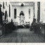 Black and white with white border photograph of the Burke Museum interior hallway with various objects, including artworks, documentation papers, iron and metal work pieces, and fabric wall hangings, placed on or against wall or in display cabinets, many of which appear to be of Asian (Chinese?) origin. 
