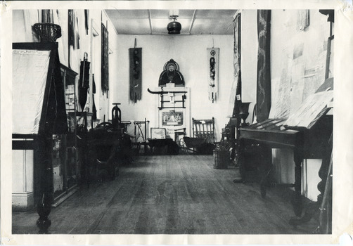 Black and white with white border photograph of the Burke Museum interior hallway with various objects, including artworks, documentation papers, iron and metal work pieces, and fabric wall hangings, placed on or against wall or in display cabinets, many of which appear to be of Asian (Chinese?) origin. 