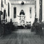 Black and white photograph of the Burke Museum interior hallway with various objects, including artworks, documentation papers, iron and metal work pieces, and fabric wall hangings, placed on or against wall or in display cabinets, many of which appear to be of Asian (Chinese?) origin. 