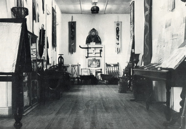 Black and white photograph of the Burke Museum interior hallway with various objects, including artworks, documentation papers, iron and metal work pieces, and fabric wall hangings, placed on or against wall or in display cabinets, many of which appear to be of Asian (Chinese?) origin. 