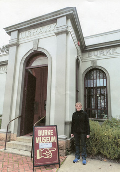 Woman wearing warm clothes is standing in front of the Burke Museum entrance. On her left side there is a sign saying "Burke Museum" with a hand pointing to the right.