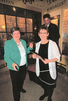 Two smiling women stand side by side, the woman on the right is holding a photo frame. Behind them stands a man and historic shopfronts.