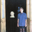 A woman stands looking at the camera, next to her is the bust of a large bald man. The woman stands in front of two french doors that are opened outwards, the bust sits within the doors.