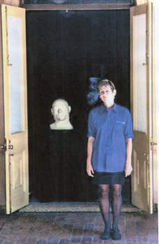 A woman stands looking at the camera, next to her is the bust of a large bald man. The woman stands in front of two french doors that are opened outwards, the bust sits within the doors.