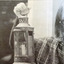 A woman stands close to the camera, she is wearing sunglasses, dangly circle earrings and a woollen shawl. She holds an antique looking lantern with a wax candle up and is peering into the camera. The photograph is in black and white.