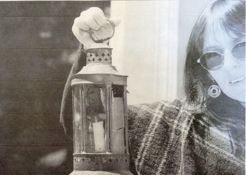 A woman stands close to the camera, she is wearing sunglasses, dangly circle earrings and a woollen shawl. She holds an antique looking lantern with a wax candle up and is peering into the camera. The photograph is in black and white.