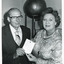 Uncropped black and white photo of man and woman smiling and holding a document up to the camera in front of museum object.