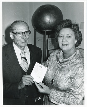 Uncropped black and white photo of man and woman smiling and holding a document up to the camera in front of museum object.