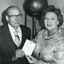 Cropped black and white photo of man and woman smiling and holding a document up to the camera in front of museum object.