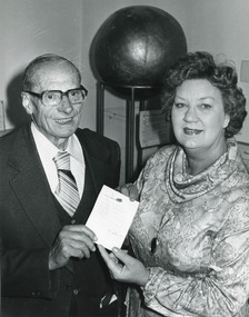 Cropped black and white photo of man and woman smiling and holding a document up to the camera in front of museum object.