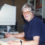 Colour photograph of woman writing into a notebook while listening to headphones, computer equipment beside her.