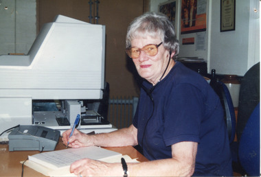 Colour photograph of woman writing into a notebook while listening to headphones, computer equipment beside her.