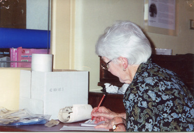 Colour photograph of woman sitting at desk, writing with a pencil.