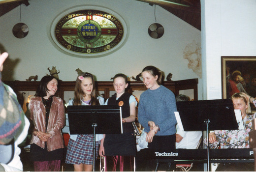 Four female singers stand behind and read from a music stand that is placed next to a seated keyboardist reading from a music stand. 