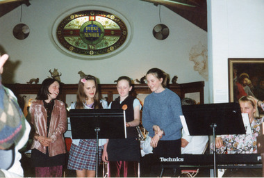 Four female singers stand behind and read from a music stand that is placed next to a seated keyboardist reading from a music stand. 