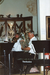 Colour photograph of two musicians seated in front of museum taxidermy mount display. The musicians wear white shirts and play brass horn instruments. A keyboard and a music stand appear on the left foreground of the image.
