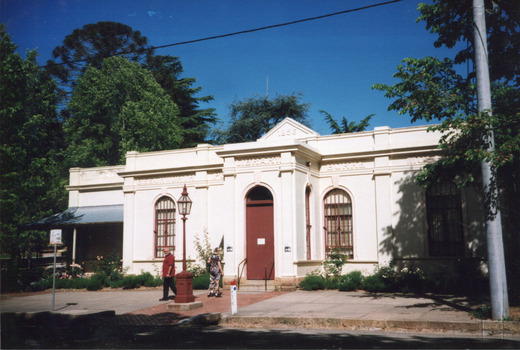 Two people stand on a footpath near a Victorian-style street lamp near the entrance to a cream stone building surrounded by green trees. The building's large red door is closed and there is a sign attached .