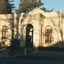 Footpath with Victorian style street lamp in front of a shaded cream stone building surrounded by trees. Two people stand in the vestibule, where the door is open.