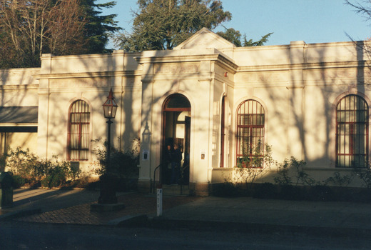 Footpath with Victorian style street lamp in front of a shaded cream stone building surrounded by trees. Two people stand in the vestibule, where the door is open.