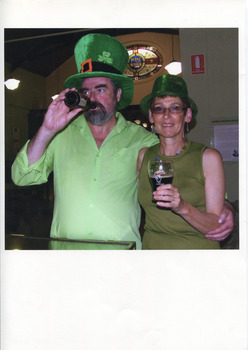 A colour photograph surrounded by a large white border. In the photograph there are two people with pale skin standing in the Burke Museum with the stained glass window in the background. Both wear green clothes and green hats. The man on the left is drinking from a beer bottle and has his arm around a woman, who is on his right holding a glass of beer.