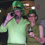 A colour photograph of two people with pale skin standing in the Burke Museum with the stained glass window in the background. Both wear green clothes and green hats. The man on the left is drinking from a beer bottle and has his arm around a woman, who is on his right holding a glass of beer.