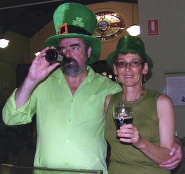 A colour photograph of two people with pale skin standing in the Burke Museum with the stained glass window in the background. Both wear green clothes and green hats. The man on the left is drinking from a beer bottle and has his arm around a woman, who is on his right holding a glass of beer.