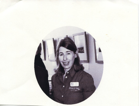 A black and white photograph in an oval shape printed on white, rectangular paper. A person with light skin and dark, shoulder-length hair is shown from the waist up, facing the camera. She wears an embroidered, button-up shirt with an Indigo Shire nametag. Behind her is a row of framed images, a display cabinet, and the back of another person