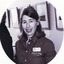 A black and white photograph in a oval shape. A person with light skin and dark, shoulder-length hair is shown from the waist up, facing the camera. She wears an embroidered, button-up shirt with an Indigo Shire nametag. Behind her is a row of framed images, a display cabinet, and the back of another person.
