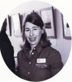 A black and white photograph in a oval shape. A person with light skin and dark, shoulder-length hair is shown from the waist up, facing the camera. She wears an embroidered, button-up shirt with an Indigo Shire nametag. Behind her is a row of framed images, a display cabinet, and the back of another person.