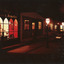 Colour photograph of  a dimly lit streetscape with a reddish hue showing a line of shops with 19th-century lamp posts. A jeweller's shop with high arched windows is in the foreground, with an image of two Victorian gentlemen visible in the window.  Source:  Burke Museum Collection.