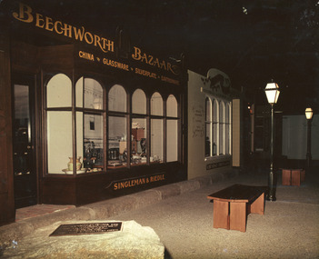 Colour  photograph featuring the shop frontage of Beechworth Bazaar with high arched windows and antique ware on display. Dimly lit streetscape lined with 19th-century lamp posts and benches, and a boulder with a plaque in the foreground. Source: Burke Museum Collection.