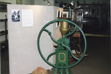 Colour photograph of grain mill as part of the Harvest Exhibition at Burke Museum in 1999. It features a grain mill, made by  B.M. Purshouse in Wolverhampton, England, with green wheels and base, and a gold container in which grain is poured. 