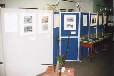 Colour photograph depicting a section of "The Harvest" exhibition with display cabinet containing objects and display boards containing photos and information.  In the foreground is believed to be an old seed distributor.