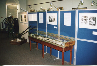 Colour photograph of "The Harvest" exhibition with a display cabinet with objects inside surrounded by blue boards displaying photographs and information. Just next to the cabinet is a seed distributor, behind which is a grain mill.
