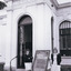 A black and white photograph taken outside the Burke Museum, in front of the door with 'Library' written on the pillar. Two light-skinned people in dresses stand around an A-frame sign with text in various fonts (see transcription). 