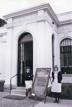 A black and white photograph taken outside the Burke Museum, in front of the door with 'Library' written on the pillar. Two light-skinned people in dresses stand around an A-frame sign with text in various fonts (see transcription). 