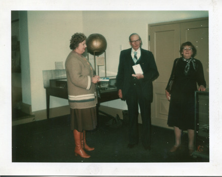 A colour photograph with a white boarder of two women and a man in front of an exhibition display table.