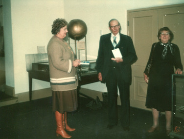A colour photograph of two women and a man in front of an exhibition display table.