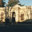 A colour photograph of the front façade of the Burke Museum, centred on the main door, stretching to one window and a shade roof on the left, and two windows on the right. The shadows of tree branches are cast along the museum façade and more trees are visible behind the museum.  Two people stand in the entryway.