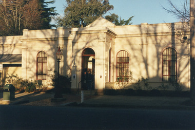 A colour photograph of the front façade of the Burke Museum, centred on the main door, stretching to one window and a shade roof on the left, and two windows on the right. The shadows of tree branches are cast along the museum façade and more trees are visible behind the museum.  Two people stand in the entryway.