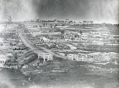 Black and white historical landscape with a small bridge in the foreground, with the road leading to the left, housing and fences to the right, and a line of trees before a hill on the horizon.