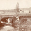 A landscape scene of a wooden bridge with a shallow creek running beneath, vegetation and rocks on either side. There are 3 children in the scene two young girls in white, one on each side of the water, the one on the right bank has a parasol. There is a boy on top of the bridge looking down at the girls. 