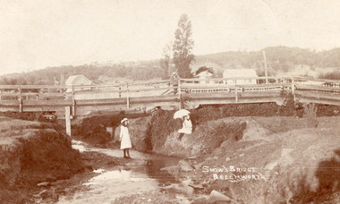 A landscape scene of a wooden bridge with a shallow creek running beneath, vegetation and rocks on either side. There are 3 children in the scene two young girls in white, one on each side of the water, the one on the right bank has a parasol. There is a boy on top of the bridge looking down at the girls. 