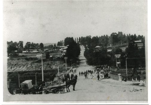 Black and white view of Camp Street and bridge over Spring Creek. Several people are on the bridge, and a horse and cart can be seen in the foreground.