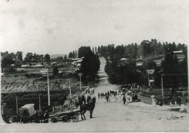 Black and white view of Camp Street and bridge over Spring Creek. Several people are on the bridge, and a horse and cart can be seen in the foreground.