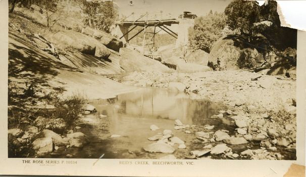 Reid's Creek, Beechworth, Sepia toned postcard with a bridge in the background and a shallow creek in the foreground. A car can be seen crossing the bridge. 