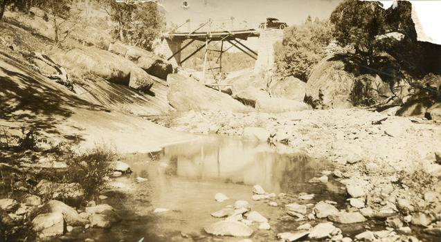 Sepia toned photograph of a creek with a bridge in the background, and a shallow creek in the foreground. A car can be seen crossing the bridge. 