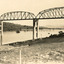 Photo is taken from the bank of a river and shows a bridge under construction with two people on a bike watching from the shore. The bridge consists of multiple iron spans on high concrete pylons. Fields are seen on the bank behind the bridge. 